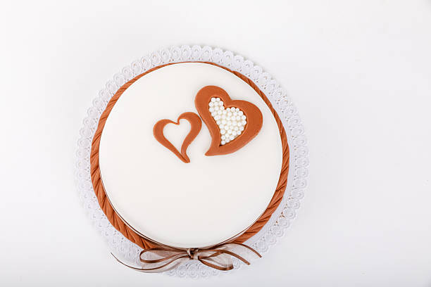 Studio shot of a frosted white cake featuring two brown heart designs on top, one filled with white candy beads. Placed on a decorative paper doily and set against a stark white background.