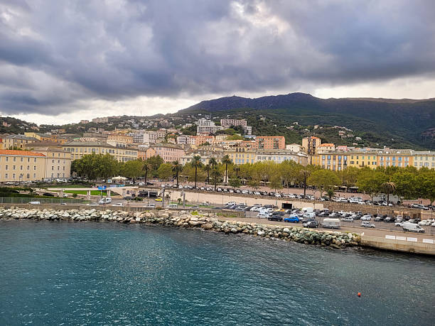 A scenic view of Bastia, Corsica, captured from a ferry approaching the harbor. The image showcases the waterfront with historic buildings leading to the main square, set against a backdrop of hills and a clear blue sky. The calm sea reflects the sunlight, creating a picturesque Mediterranean atmosphere. Perfect for themes of travel, tourism, coastal beauty, and cultural heritage.