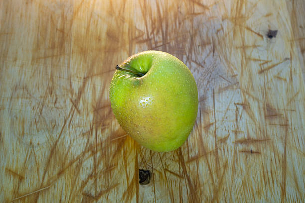 A single fresh green apple placed on a scratched wooden surface. The apple appears crisp and slightly shiny, with visible water droplets that emphasize freshness. The rustic wooden background adds texture and contrast, creating a natural and organic aesthetic. Ideal for themes related to healthy eating, nutrition, simplicity, and farm-to-table concepts.