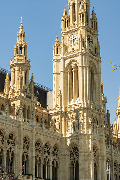The image showcases the magnificent Vienna City Hall (Wiener Rathaus), an iconic landmark and architectural masterpiece in Austria. The building features a stunning Neo-Gothic design with ornate stonework, pointed arches, and intricate detailing. Dominating the scene is the tall central clock tower, flanked by smaller spires and decorative statues. The light-colored stone façade contrasts beautifully with the clear blue sky, highlighting the elegance and grandeur of this historic structure. This landmark is a symbol of Vienna’s cultural heritage and 19th-century European architecture.