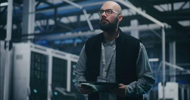 istock Bearded IT Engineer in Dark Vest and Glasses Monitors System Metrics on Digital Tablet 2253677741