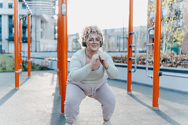 Plus size woman exercising with squats at an outdoor gym, listening to music on headphones