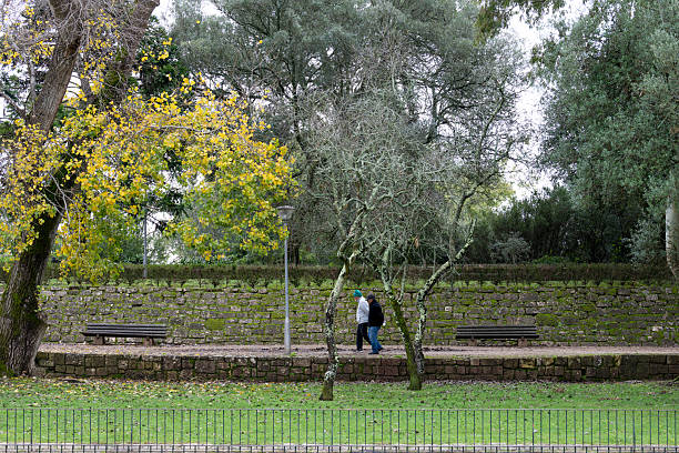 Lisbon Portugal December 07 2025. Two people walk along a stone path with benches and trees inside Monsanto Forest Park.