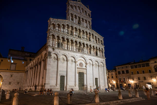 26 July 2025 Lucca, Italy. Night view of an illuminated Romanesque church with layered arches and ornate stone façade, surrounded by a lively historic square, capturing atmosphere, heritage, and architectural detail at Chiesa di San Michele in Foro, Lucca, Italy.