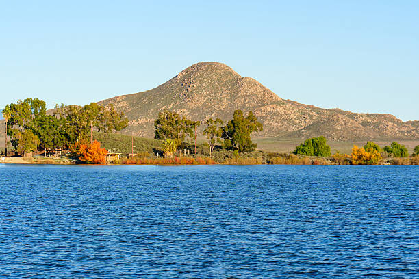 Blue waters of Lake Perris extend to a tree-lined shore with green and vibrant autumn foliage, backed by a rugged mountain under a clear, bright sky.