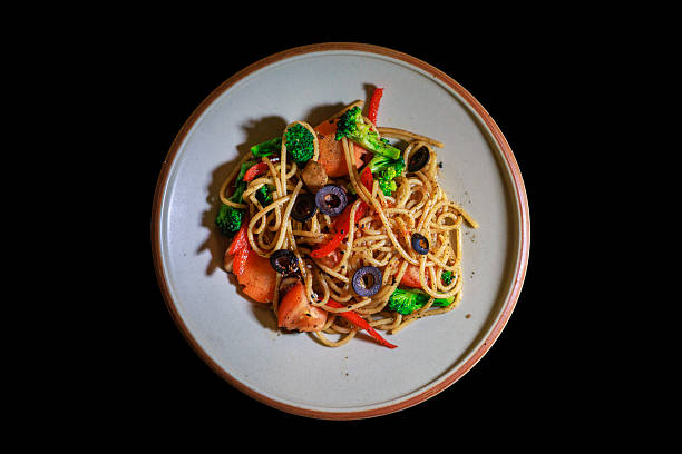 Close-up of vegetarian aglio olio pasta prepared with spaghetti, broccoli, red bell pepper, tomatoes, and black olives. A simple plant-based Italian dish served on a ceramic plate, highlighting fresh ingredients, olive oil seasoning, and comfort food dining.