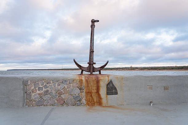 A rusty anchor sculpture stands on a concrete wall by the water, set against a serene cloudy sky. The anchor's weathered texture and the stone wall's rustic charm evoke a sense of stability and hope.