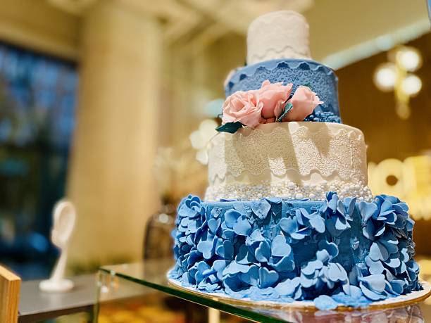 Three Tier Blue Floral Cake Showcased On Glass Stand, Adorned With Fondant Hydrangeas And Sugar Roses, Lace Texture On Buttercream Tiers, Soft Pastel Blue And Ivory Palette, Ambient Ballroom