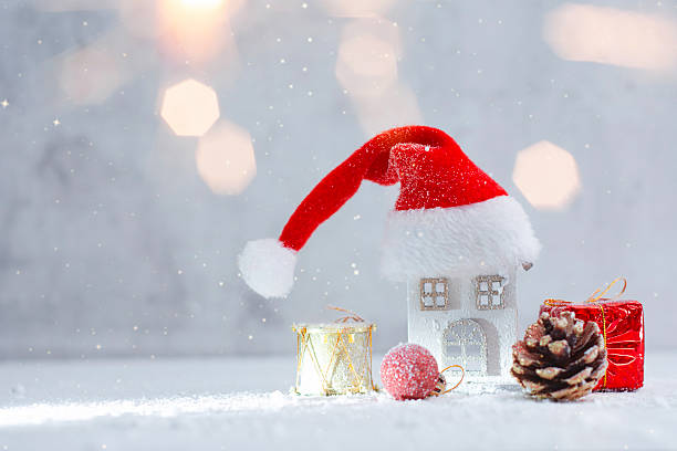 istock Small decorative Christmas house wearing a Santa hat surrounded by a gift box, pine cone and ornaments in snow. Festive winter holiday concept for Christmas greeting cards and New Year marketing. 2253727532