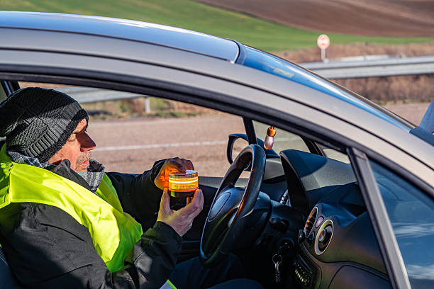 Driver in the foreground placing a V16 warning or emergency beacon with a broken-down vehicle on the road, pre-signaling. Beacon mandatory in Spain by 2026. Tordesillas, Valladolid, Castile and León, Spain, Europe - December 21, 2025.