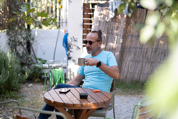 Man Sitting at Garden Table Drinking Hot Beverage. Morning Coffee or Tea in the Garden. Man Having a Warm Drink at Outdoor Table. Relaxed Man Enjoying Hot Drink Outdoors.