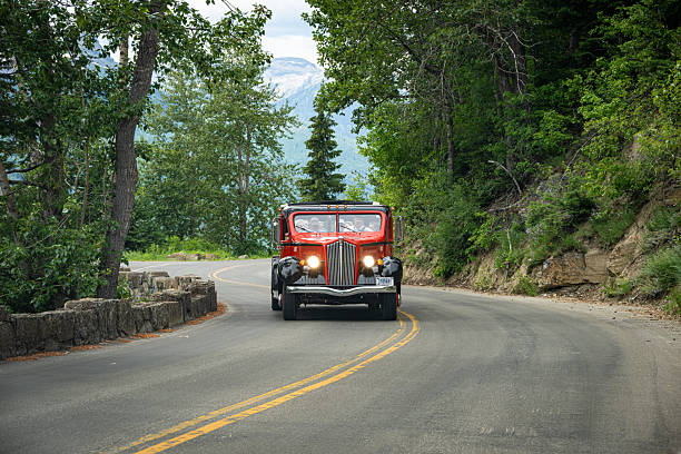 Tourists riding a historic red tour bus along Going-to-the-Sun Road in Glacier National Park, Montana, USA, during summer. Scenic alpine landscape with steep green slopes, rocky cliffs, and wilderness surroundings, illustrating tourism and sightseeing in a national park.