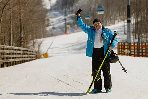 Active senior man with skis and winter equipment standing outdoors at a ski resort, using smartphone and enjoying winter vacation