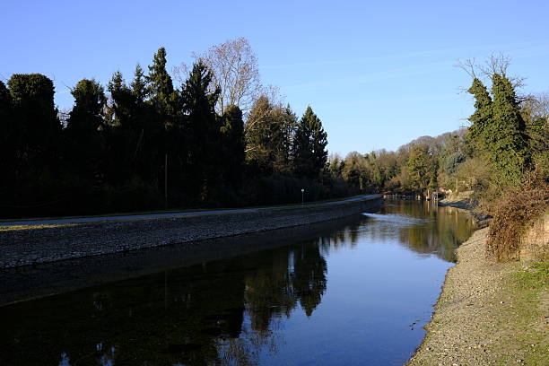 Peaceful river canal flowing through a natural landscape, with trees reflected on the calm water under a clear blue winter sky. Stone embankments and woodland surroundings create a serene outdoor scene, ideal for themes of nature, tranquility, environment, and slow living.