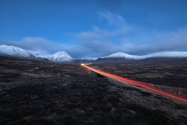 Long exposure view of light trails on a remote road crossing a dark mountain landscape at dusk, with snow-capped peaks and dramatic sky creating a sense of motion and solitude. Scottish Highlands.