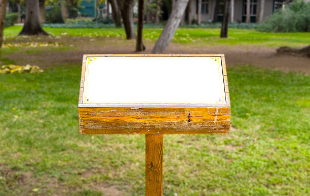 istock Editable wooden signpost with blank white display—framed by green grass, trees, and distant buildings in a tranquil park or garden setting under clear daylight. 2253759602
