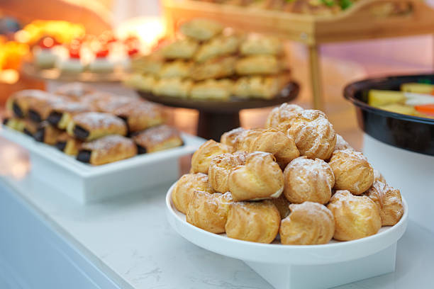 Elegant dessert table featuring a plate of golden cream puffs alongside assorted pastries and sweets displayed on tiered stands. Styled for a celebration or event setting, highlighting modern dessert presentation, hospitality, and indulgent food styling indoors.