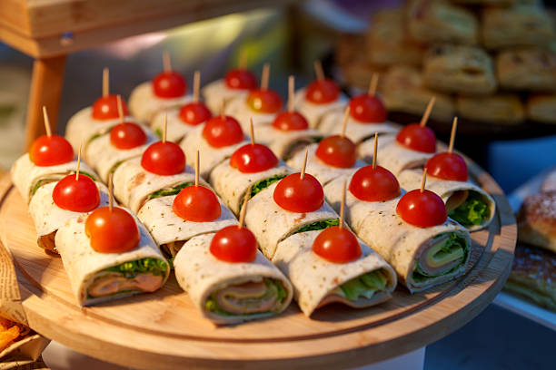 Catering platter of assorted wrap sandwiches topped with cherry tomatoes and secured with wooden picks, arranged on a wooden serving board. Ideal for buffet, party, or corporate event settings, showcasing fresh ingredients, finger food presentation, and modern hospitality dining indoors.