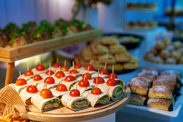 Catering platter of assorted wrap sandwiches topped with cherry tomatoes and secured with wooden picks, arranged on a wooden serving board. Ideal for buffet, party, or corporate event settings, showcasing fresh ingredients, finger food presentation, and modern hospitality dining indoors.