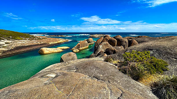 A panoramic view showcases a coastal landscape with turquoise waters, rugged rocks, and a clear blue sky, highlighting nature's beauty.