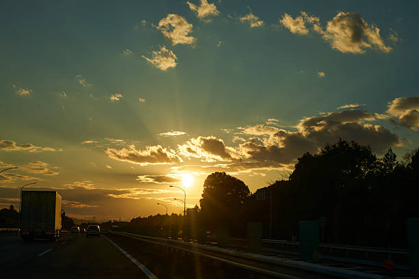 The sinking sun casts streaks of light through clouds, gently enveloping cars on the highway. A moment where the day's end meets the excitement of returning to a place of rest.