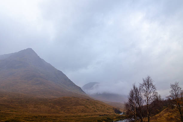 High peaks loom in a gray sky, enveloped by swirling clouds and mist. The rugged terrain below hints at Scotland's wild beauty on a chilly, overcast day.