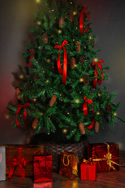 istock Christmas tree with a large red bow. The tree is decorated with cones, red satin bows and a garland. The tree flashes yellow lights. Under the Christmas tree New Year gifts. Vertical photo banner copy space 2253773197