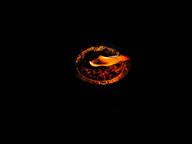 close-up view of a diya (small oil lamp) floating on dark water. The diya is housed in a natural, biodegradable bowl made of leaves, filled with orange and red flower petals, and a single bright flame at the center.