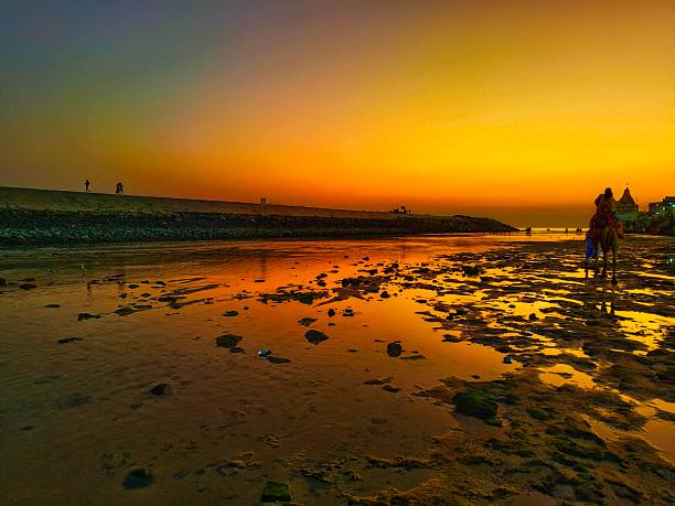 Photograph captures the stunning golden hour at a beach, likely in Gujarat, India. The setting sun paints the sky and the wet, reflective sand in vibrant hues of orange and yellow, with a deep green and blue gradient near the top