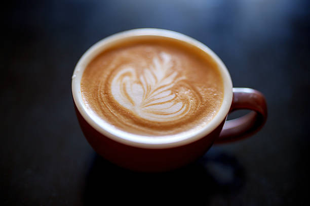 Close-up, high angle view of a latte coffee featuring delicate milk foam art served in a ceramic cup against a black background.
