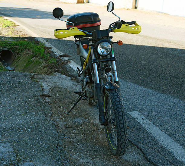 Compact yellow motorcycle with handguards rests on a cracked curb beside a road. Top case and round headlamp suggest urban commuting and light adventure.