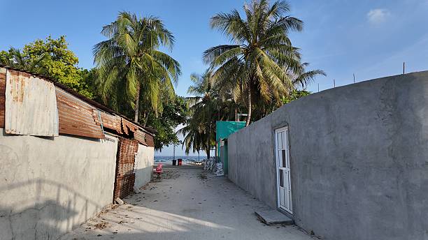A sandy path winds through rustic buildings towards the tropical ocean under a clear blue sky.