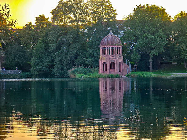 Scenic view of a historic rotunda or gazebo on the shore of a calm lake during golden hour. The elegant architectural structure is perfectly reflected in the still water, surrounded by lush green trees.