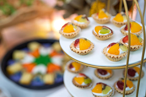 Assorted mini fruit tarts topped with fresh mango, kiwi, strawberry, and cream are neatly arranged on an elegant tiered dessert stand.