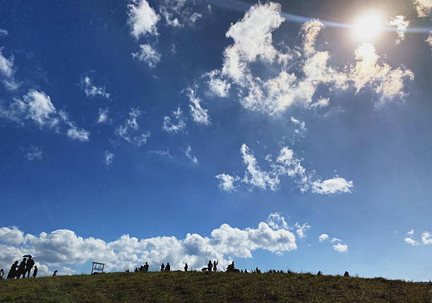 A bright blue sky with fluffy white clouds and a shining sun hangs over a grassy hill. The silhouettes of several people are visible.