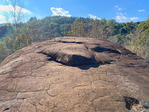 istock A close-up image of a large rock with natural cracks all over its surface, against a backdrop of lush hills and a bright blue sky. 2253786751