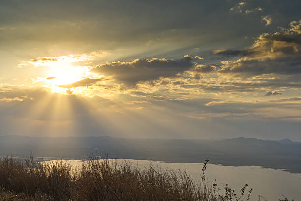 Breathtaking golden hour landscape featuring sun rays piercing through clouds over a serene reservoir. Misty mountains and silhouetted grass create a tranquil, majestic atmosphere.