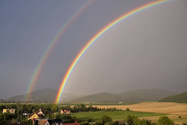 Wide-angle view of a vibrant double rainbow arching over a picturesque landscape of rural village houses, green fields, and rolling hills.