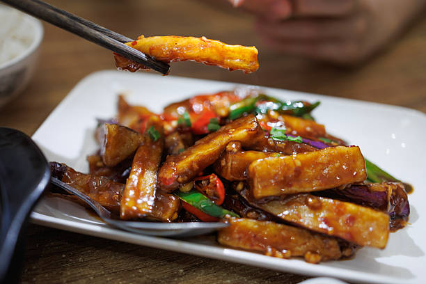 Close-up of stir-fried eggplant coated in spicy chili sauce, served on a white plate and picked up with chopsticks. This flavorful Asian vegetable dish features tender eggplant, chili peppers, and savory sauce, commonly enjoyed as a popular Chinese cuisine comfort food in restaurants.