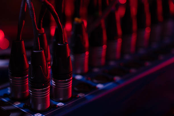 A musician in a warm recording studio holds a coiled cable while a connector is in focus, with drums and acoustic panels creating a creative audio engineering atmosphere.