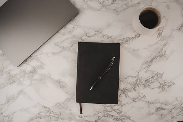istock A simple workspace features a dark notebook and pen beside a laptop on a marble table. A white coffee cup with black coffee adds warmth to this organized setup, ideal for productivity 2253813335