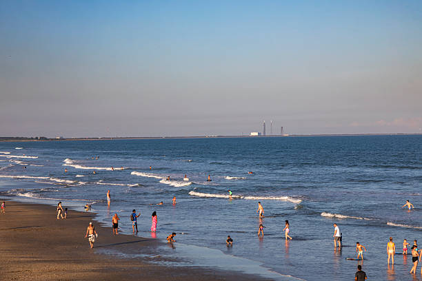 People of all ages wade and play in the shallow waters along the shoreline at Cocoa Beach, Florida, as gentle waves roll in from the Atlantic Ocean. Captured during late afternoon light, the scene conveys a relaxed coastal atmosphere with families, swimmers, and surfers spread across the water and wet sand. In the distance, the flat horizon and faint industrial structures of Florida’s Space Coast add subtle geographic context.