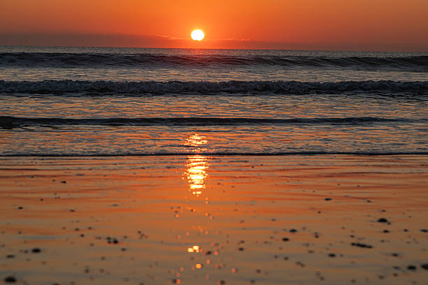 Golden sunrise over the Atlantic Ocean at Cocoa Beach, Florida, with the sun emerging above the horizon and reflecting across wet sand and gentle ocean waves. The warm orange and amber tones of the sky contrast with the cool blues of the water, creating a calm and peaceful coastal atmosphere.