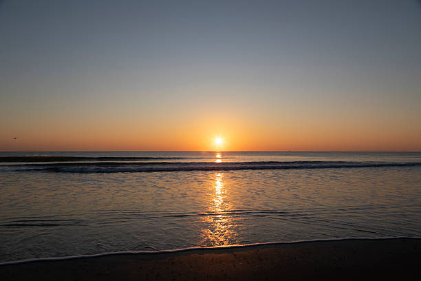 Golden sunrise over the Atlantic Ocean at Cocoa Beach, Florida, with gentle waves rolling onto dark, wet sand. The rising sun creates a warm reflection across the water, contrasting with the cool blue tones of the early morning sky.