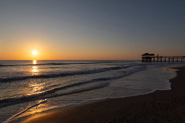 A serene sunrise unfolds over Cocoa Beach, Florida, as the first light of day warms the Atlantic Ocean and paints the sky in soft gradients of gold, orange, and blue. Gentle waves roll toward the shoreline while the iconic pier stretches into the sea, silhouetted against the glowing horizon.