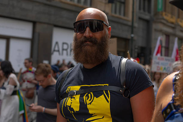 bearded man at pride parade smiling, wearing sunglasses and graphic shirt, backpack straps visible, standing among crowd with banners and rainbow flags, candid CZ, Prague, Karlova, 5.8.25