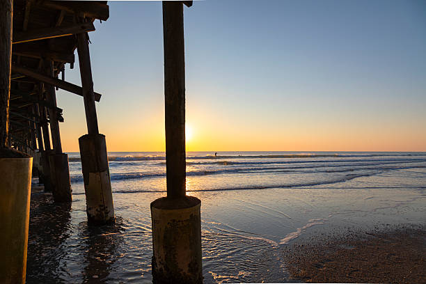 Early morning sunlight rises over the Atlantic Ocean at Cocoa Beach, Florida, casting a warm golden glow beneath a wooden pier. The sun reflects on wet sand and shallow surf, creating dramatic highlights and elongated shadows among the pier’s pilings.