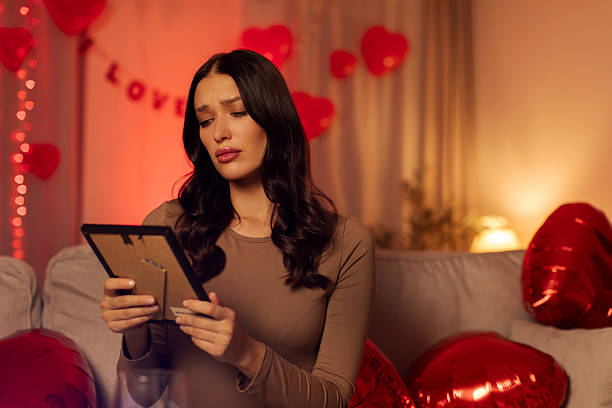 istock Woman sitting on sofa looking sad while holding picture frame, feeling lonely on Valentine's day. Room decorated with red hearts and soft lights 2253820447