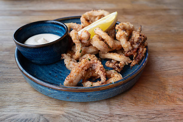 istock Crispy squid calamari, deep fried and dusted with salt and pepper alongside a bowl of herb aioli. The food is in a blue dish and on a wooden table. 2253821917