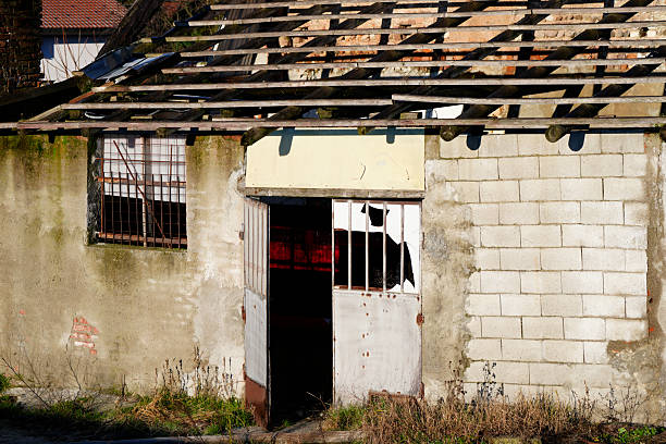 Abandoned rural building with a partially collapsed roof, weathered walls, and a broken metal door. Decay and deterioration visible in an old countryside structure, photographed in natural daylight with no people.